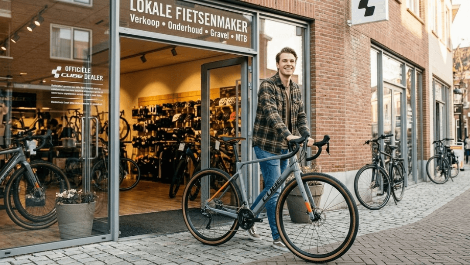 Een lachende, beginnende fietser loopt met een gloednieuwe Cube gravelbike aan de hand de lokale fietsenwinkel uit.