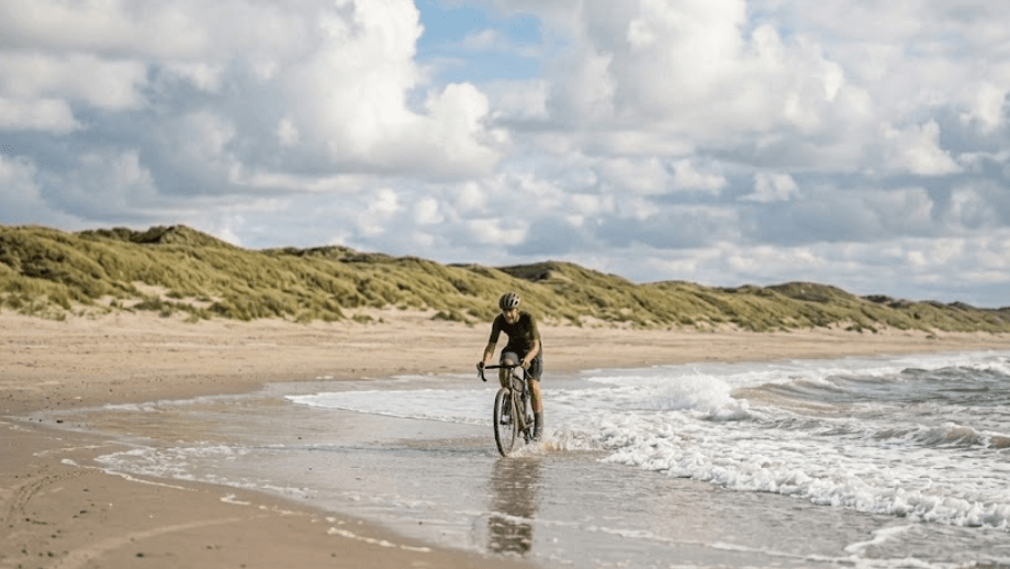 Een gravelbiker fietst vlak langs de branding op een breed zandstrand met duinen op de achtergrond, een unieke ervaring op de mooiste gravelroutes in Nederland.