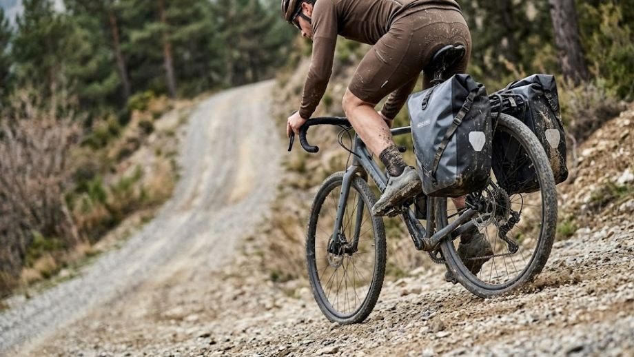 Gravelbiker klimt op de pedalen waarbij de hiel dicht bij de achterste fietstas komt, gemonteerd via de achterste bevestigingspunten gravelbike.